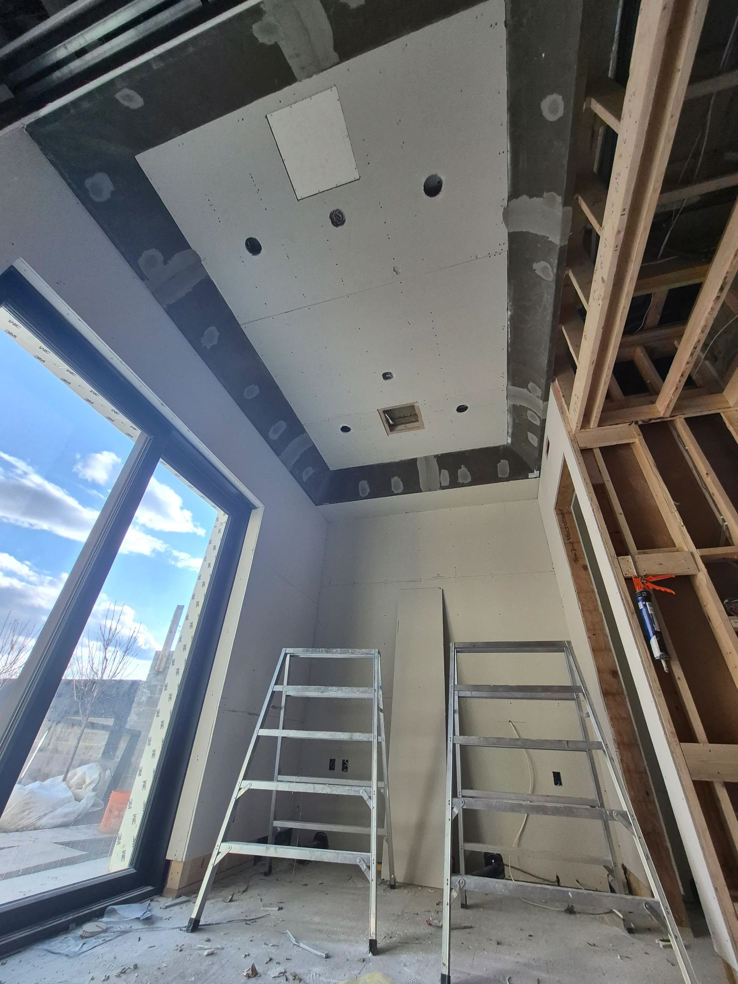 Interior view of a room under construction showing exposed wooden framing, insulation, wiring, and an unfinished ceiling with a window on the left side.