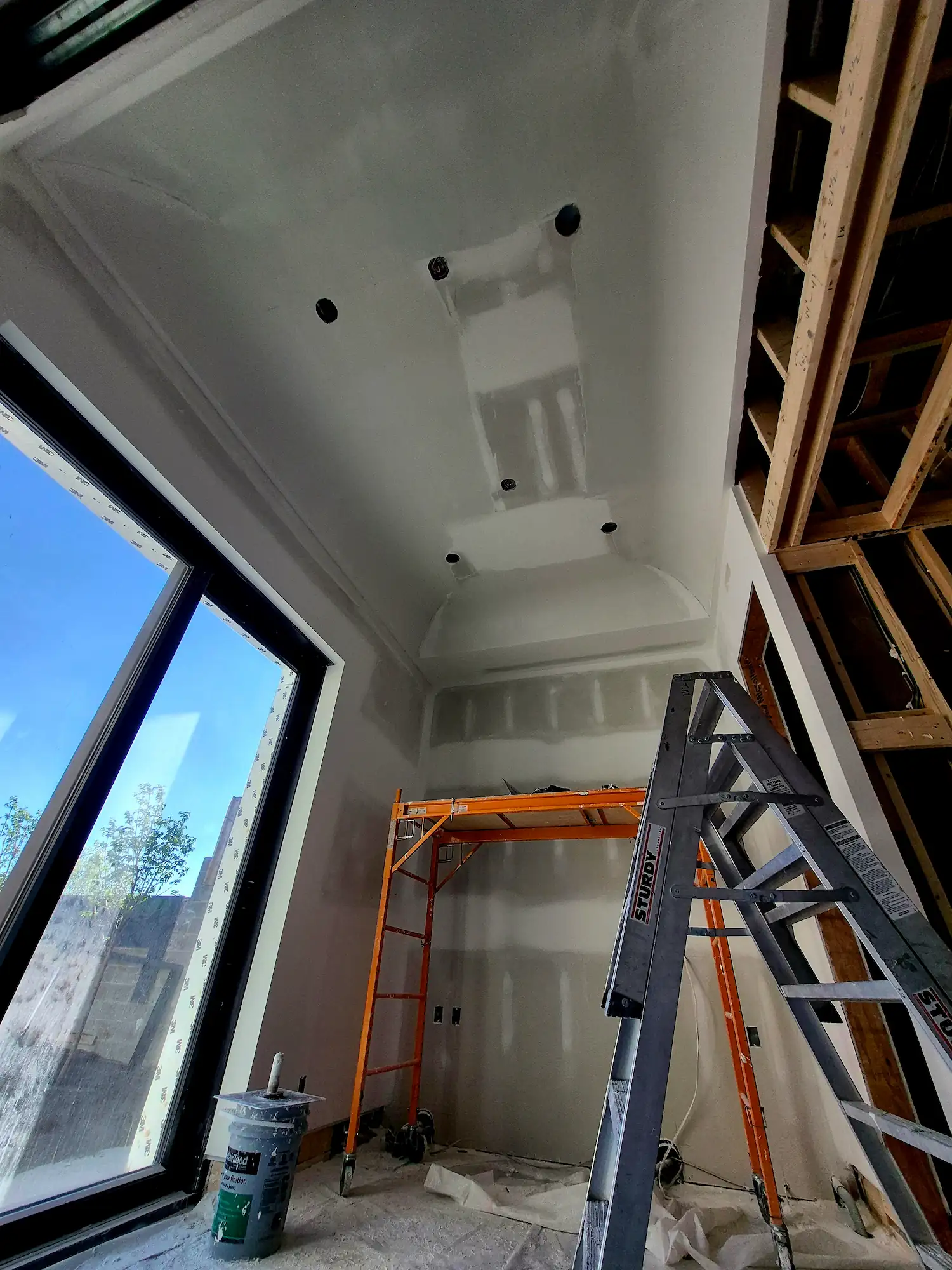 Indoor construction site with drywall installation, two metal step ladders, exposed wooden framing, and large window showing cloudy sky.