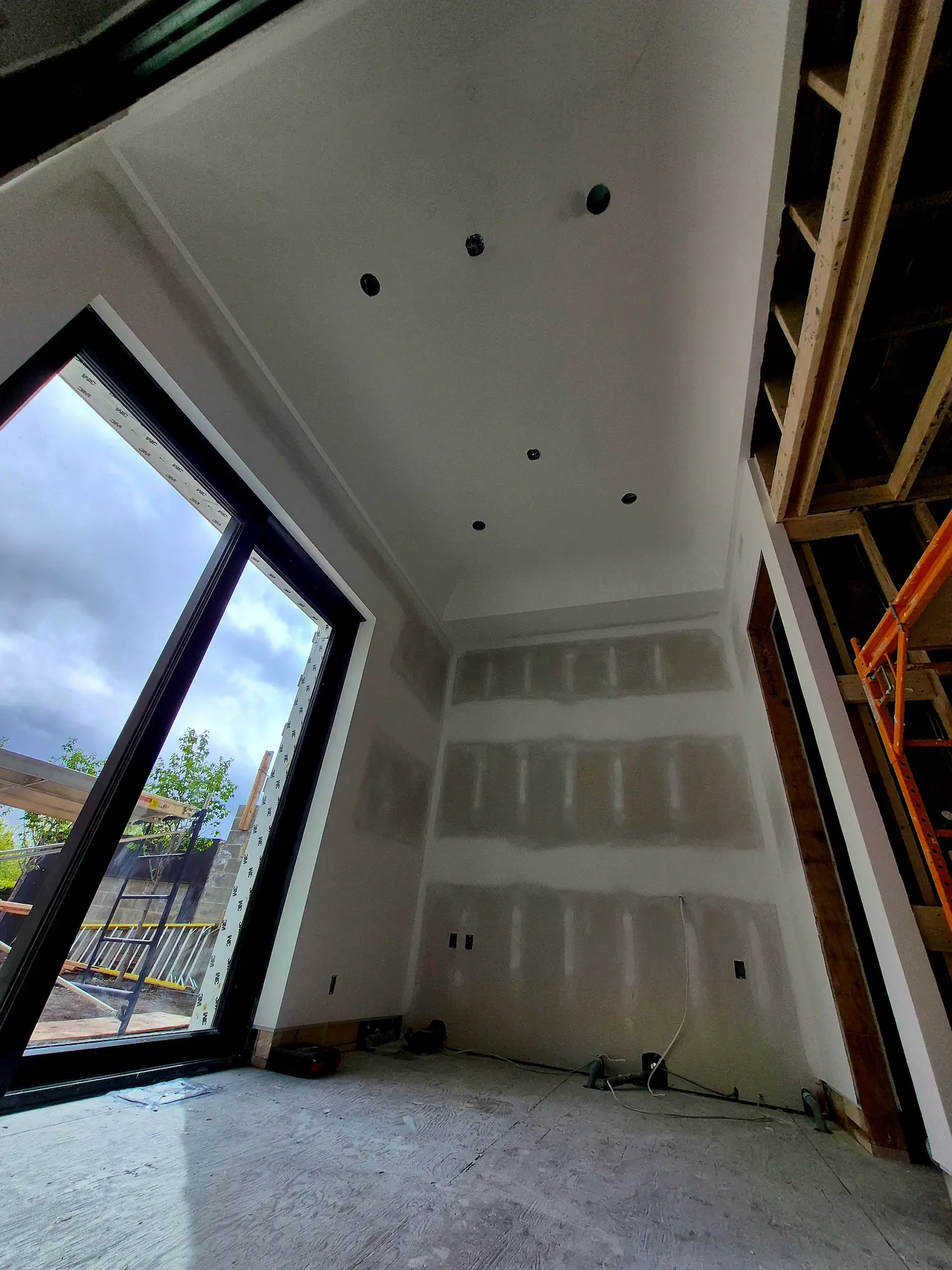 Indoor construction site showing unfinished drywall with ladder, large window, and exposed wooden framing on ceiling and wall.