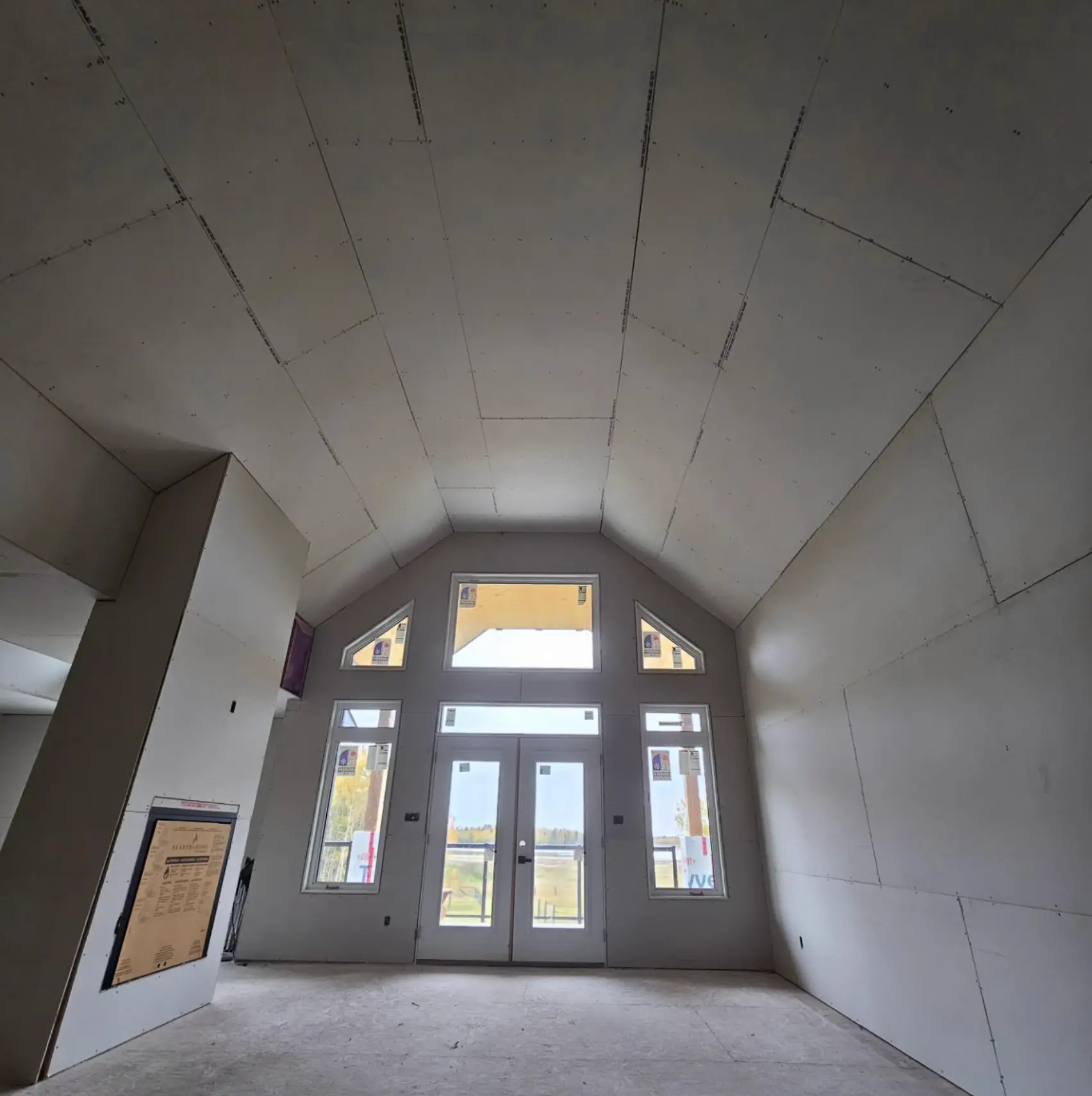 Empty room under construction with high vaulted ceiling, drywall patches, and double glass doors with sidelights and transom windows showing outdoor landscape.
