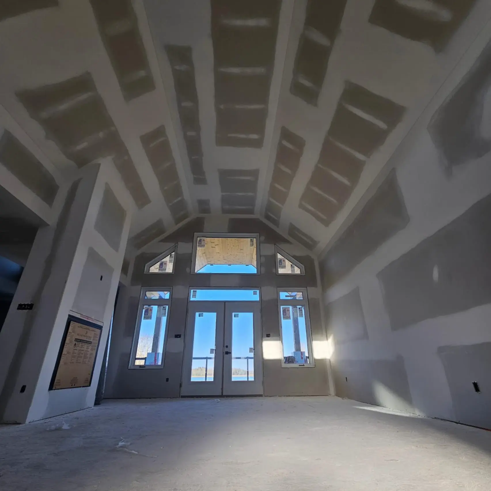 Interior of a large room under construction with drywall installed and seams taped, high vaulted ceiling, front door, and construction tools scattered around.